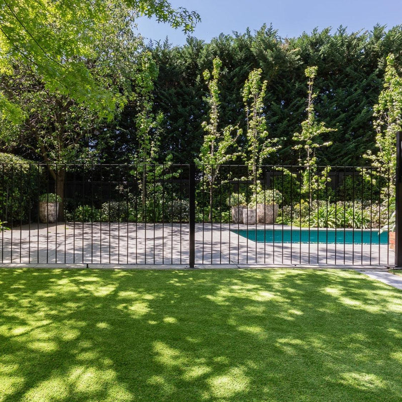 Synthetic grass backyard with pool, greenery, and black metal fence in residential setting