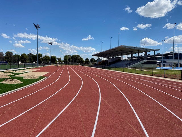 Red synthetic athletics running track with grandstand, blue sky, and green sports field